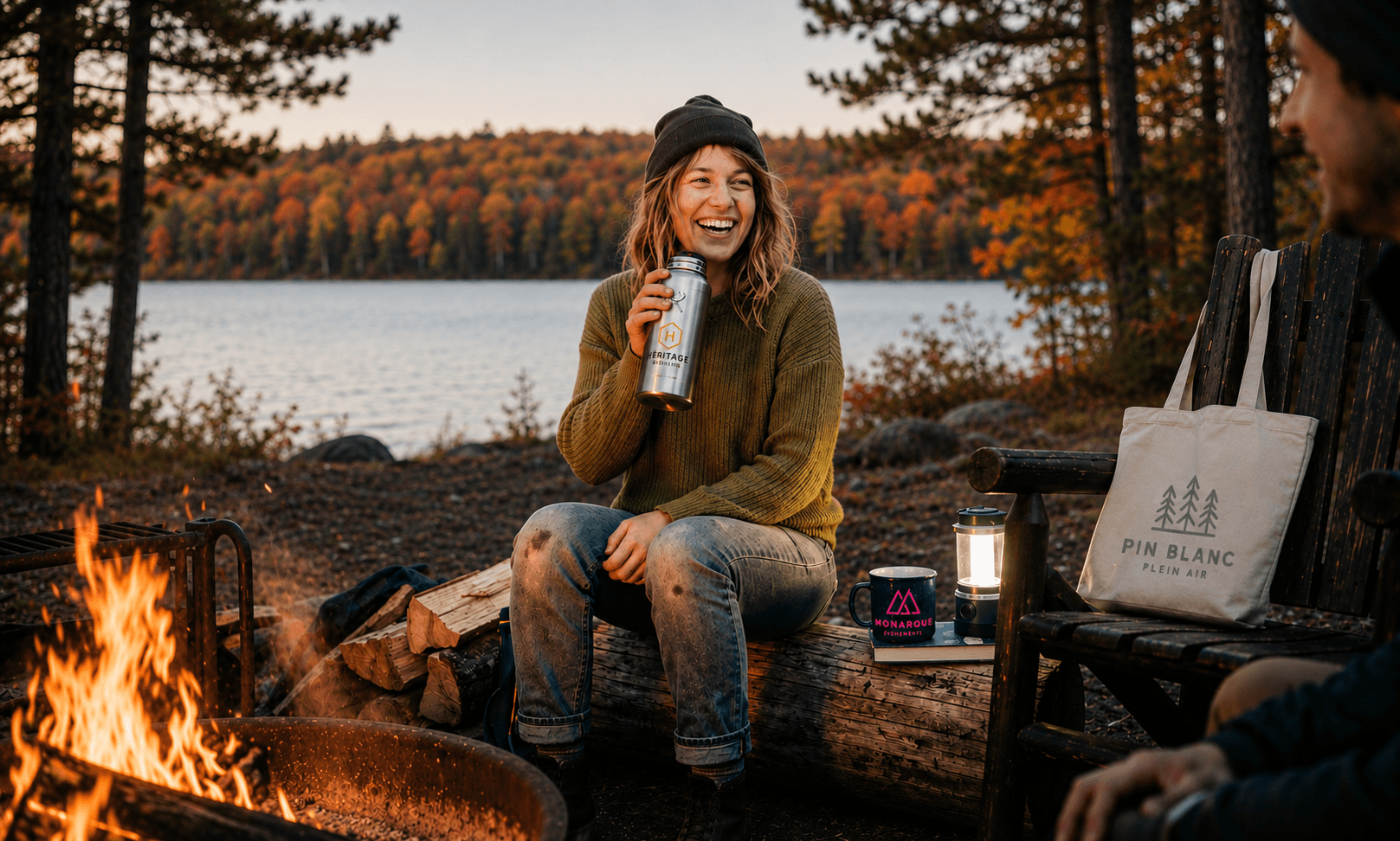 Personne assise près d'un feu de camp au bord d'un lac, tenant une bouteille isotherme personnalisée, avec une tasse et un sac fourre-tout logotés à proximité